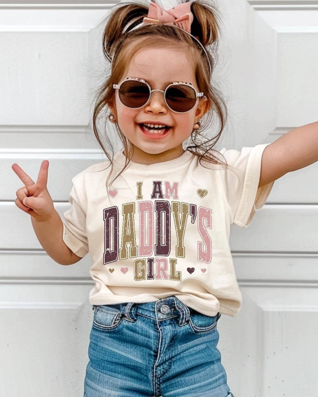 Child wearing a 'I am daddy's girl' t-shirt and denim shorts standing in front of a white garage door.