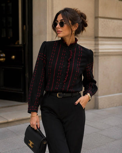 A woman wearing a black long-sleeve blouse with red vertical stripe patterns and red ruffle trim, paired with black trousers, sunglasses, and holding a black handbag with gold hardware.