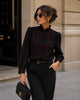 A woman wearing a black long-sleeve blouse with red vertical stripe patterns and red ruffle trim, paired with black trousers, sunglasses, and holding a black handbag with gold hardware.