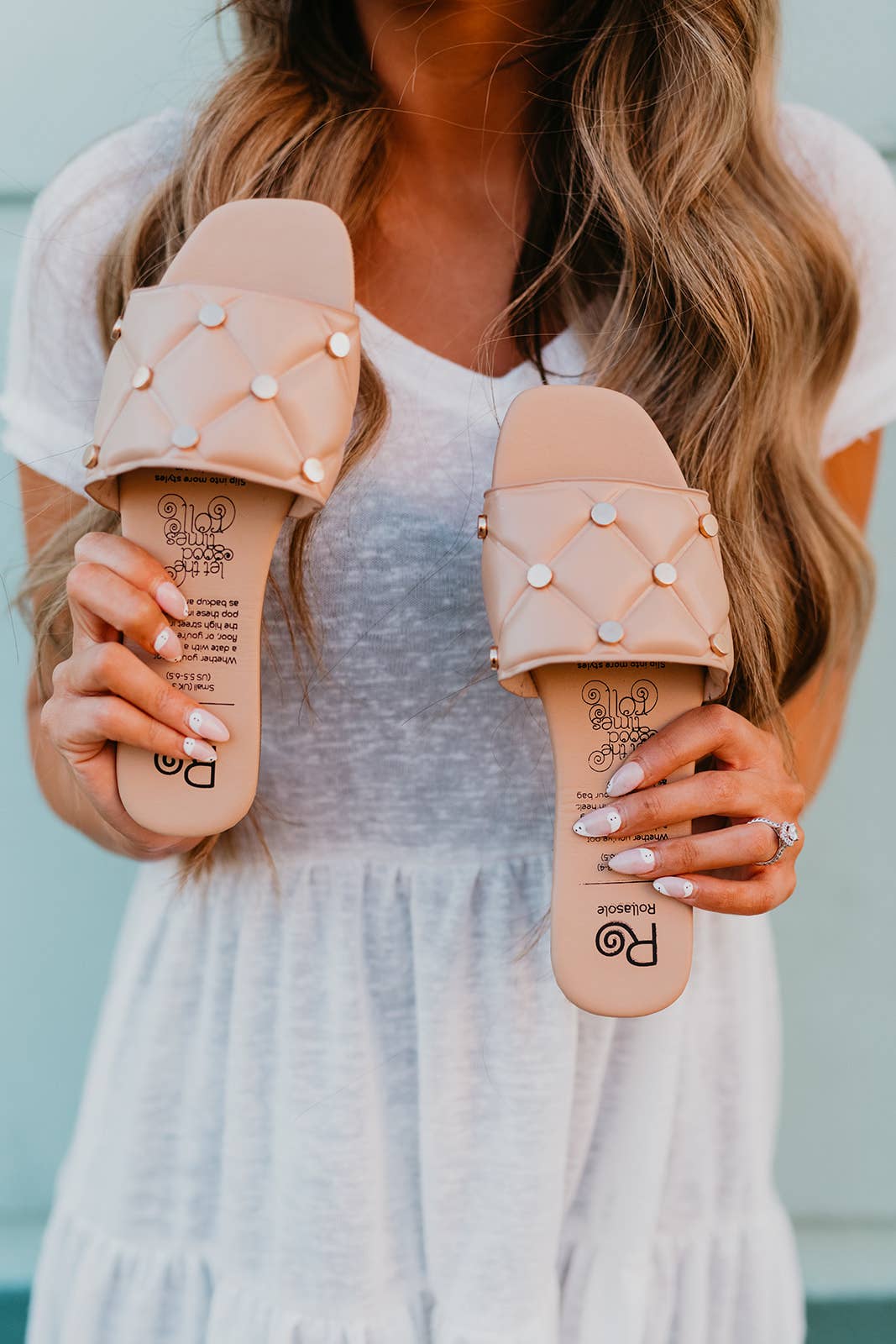 Person holding two pairs of beige sandals with decorative elements against a blurred background