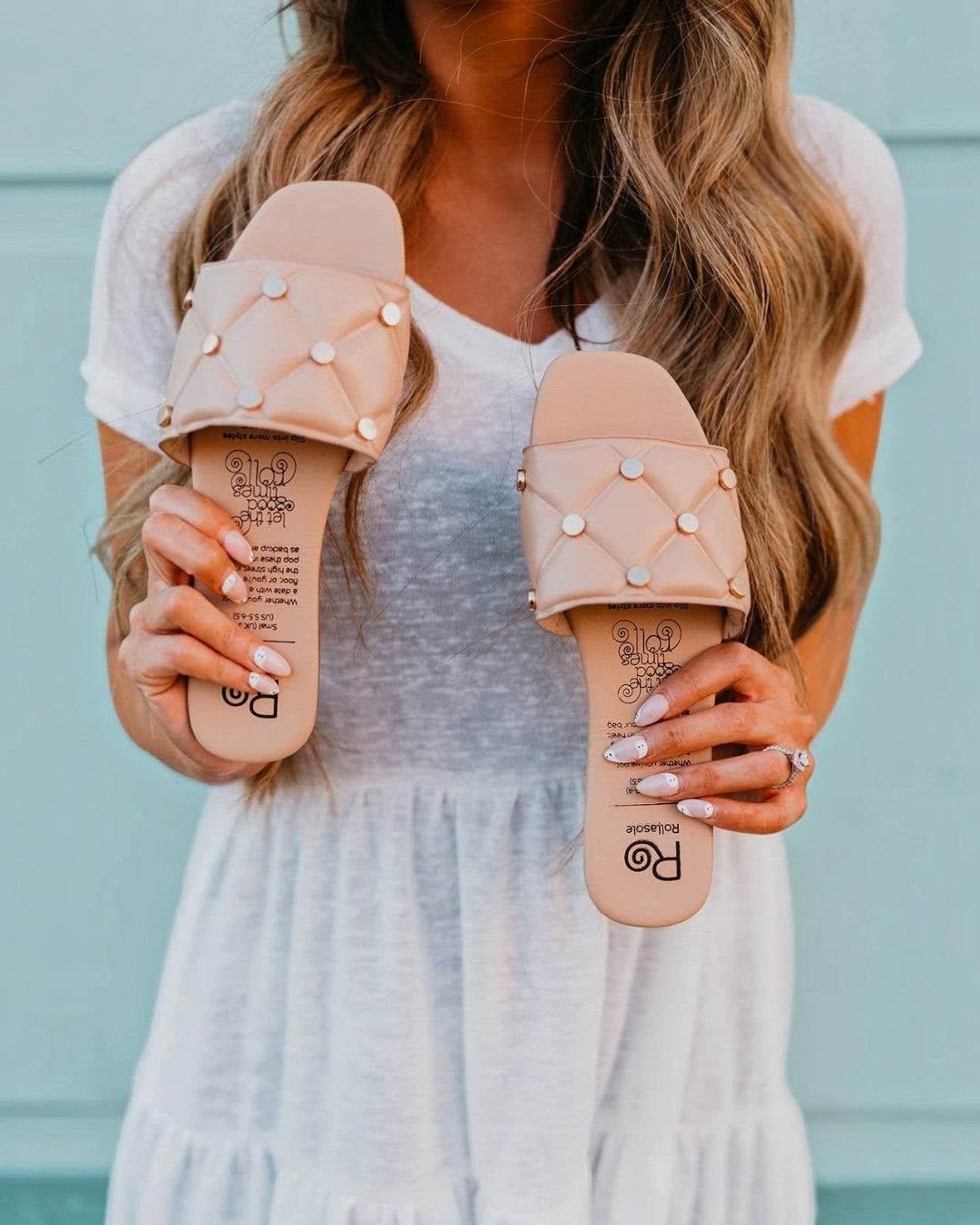 Person holding two pairs of beige sandals with decorative elements against a blurred background