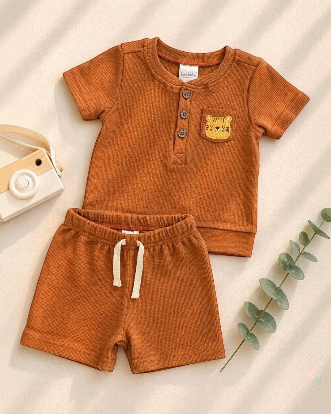 Brown children's outfit with shirt and shorts on a beige background, accompanied by a toy camera and wooden giraffe.