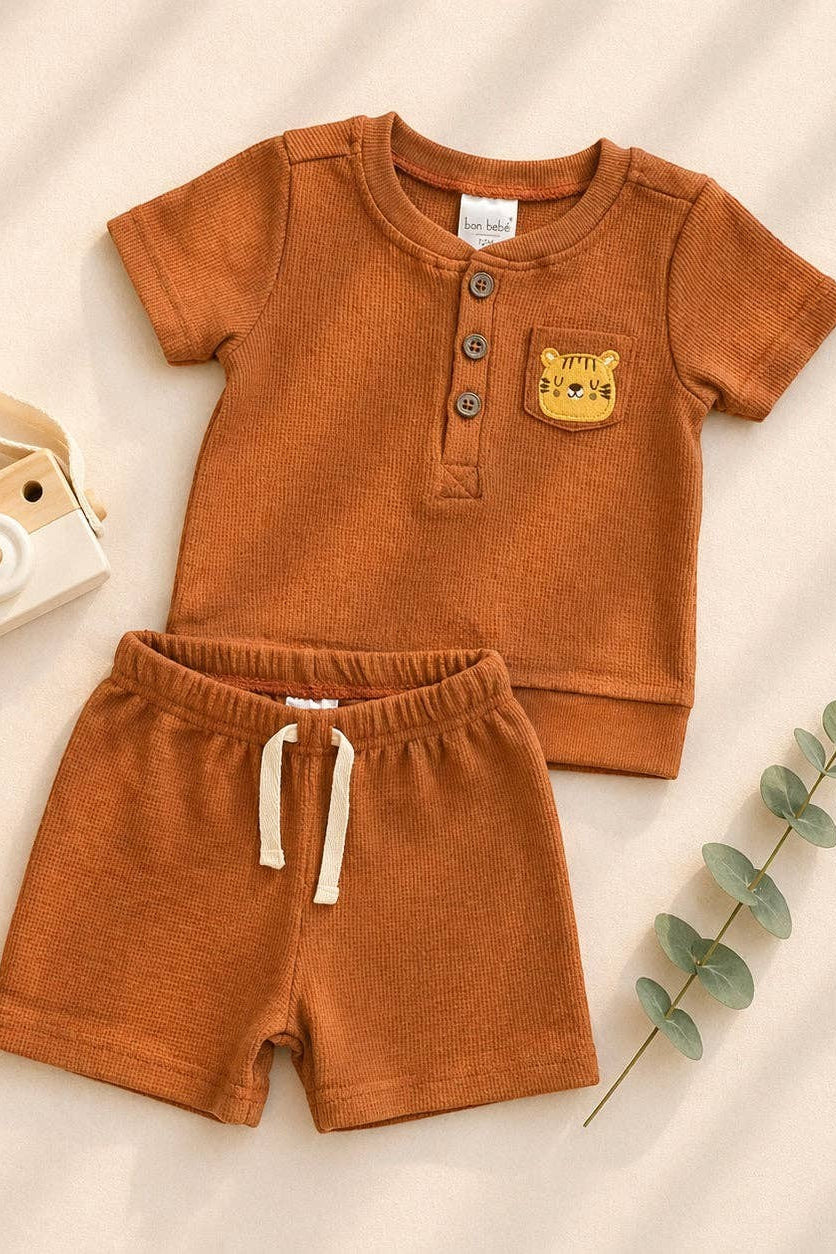 Brown children's outfit with shirt and shorts on a beige background, accompanied by a toy camera and wooden giraffe.