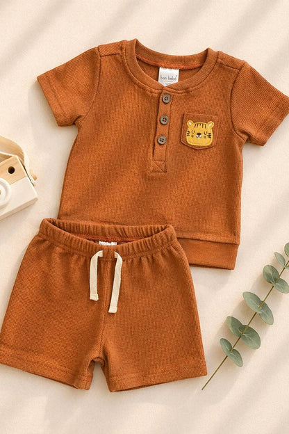 Brown children's outfit with shirt and shorts on a beige background, accompanied by a toy camera and wooden giraffe.