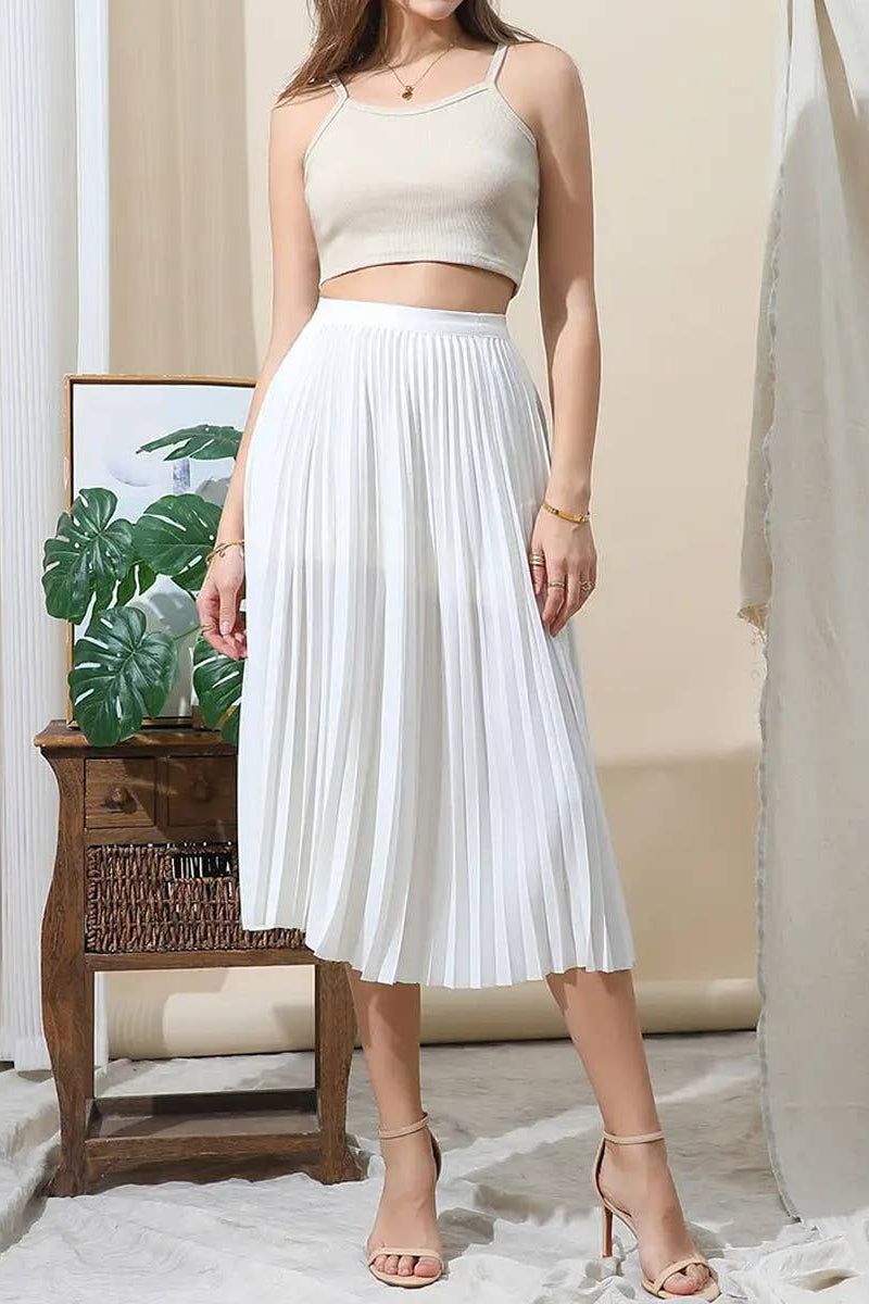 Woman wearing a white pleated skirt and beige top in a room with a plant and wooden side table.