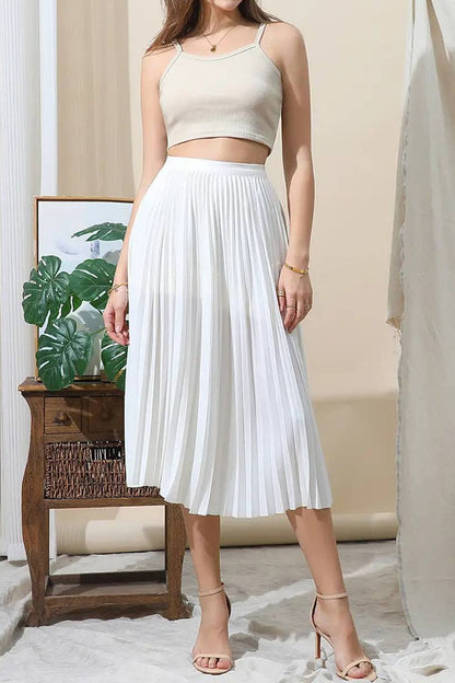Woman wearing a white pleated skirt and beige top in a room with a plant and wooden side table.