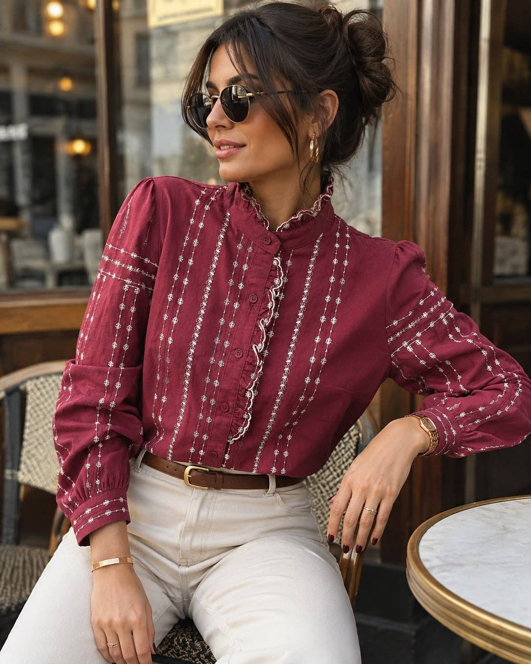 A woman wearing a burgundy long-sleeve blouse with white vertical embroidered stripes and ruffle trim, paired with cream pants, sitting at an outdoor cafe table.