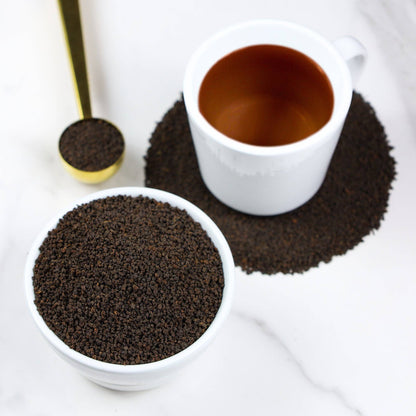 A white ceramic cup filled with a dark brown liquid sits on a bed of dark brown granules, with a matching white bowl of the same granules in the foreground and a gold spoon holding some granules on a light marble surface.