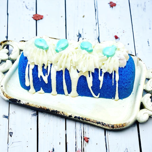 A blue twinkie bath bomb topped with white frosting and teal sugar sprinkles on a white tray against a wood background