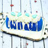 A blue twinkie bath bomb topped with white frosting and teal sugar sprinkles on a white tray against a wood background