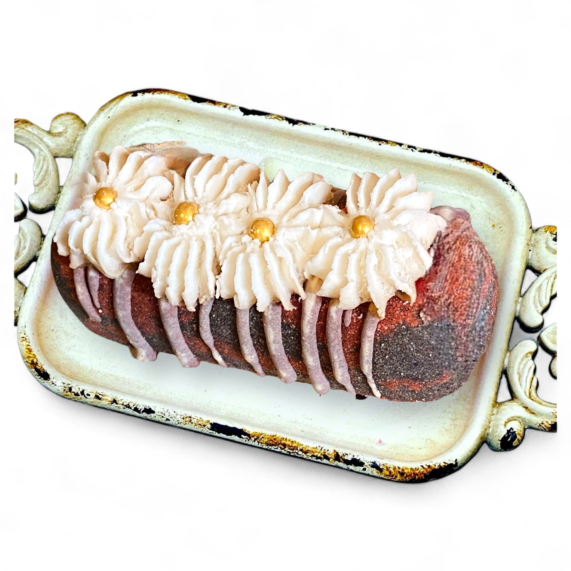 A red and black twinkie bath bomb topped with white frosting and gold sugar sprinkles on a white tray against a white background