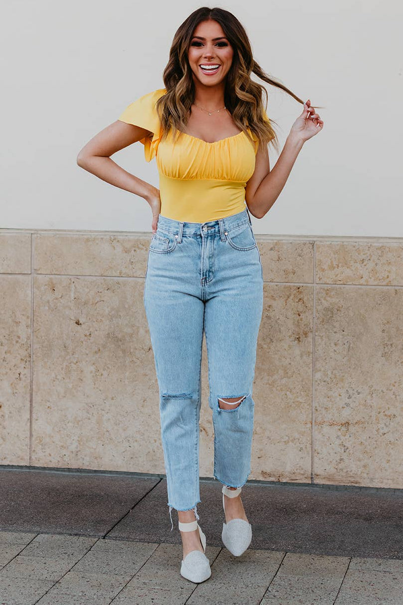 Woman in yellow top and light blue jeans standing against a beige wall.