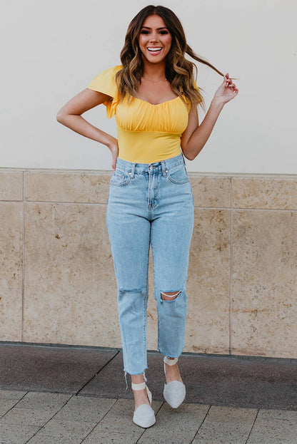 Woman in yellow top and light blue jeans standing against a beige wall.