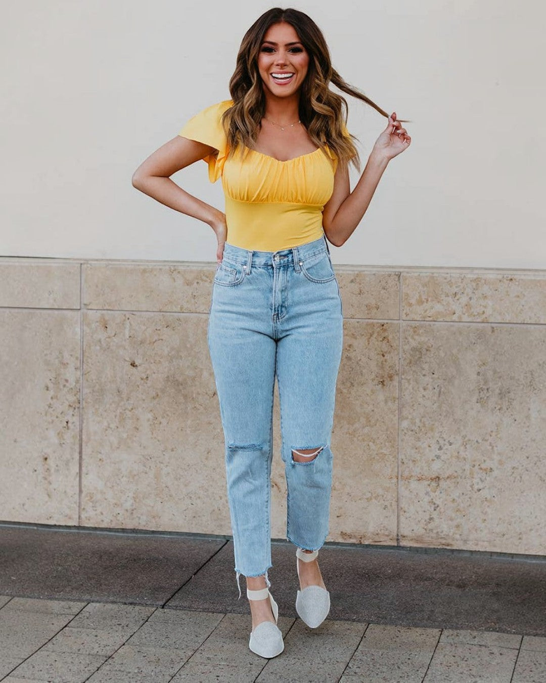 Woman in yellow top and light blue jeans standing against a beige wall.