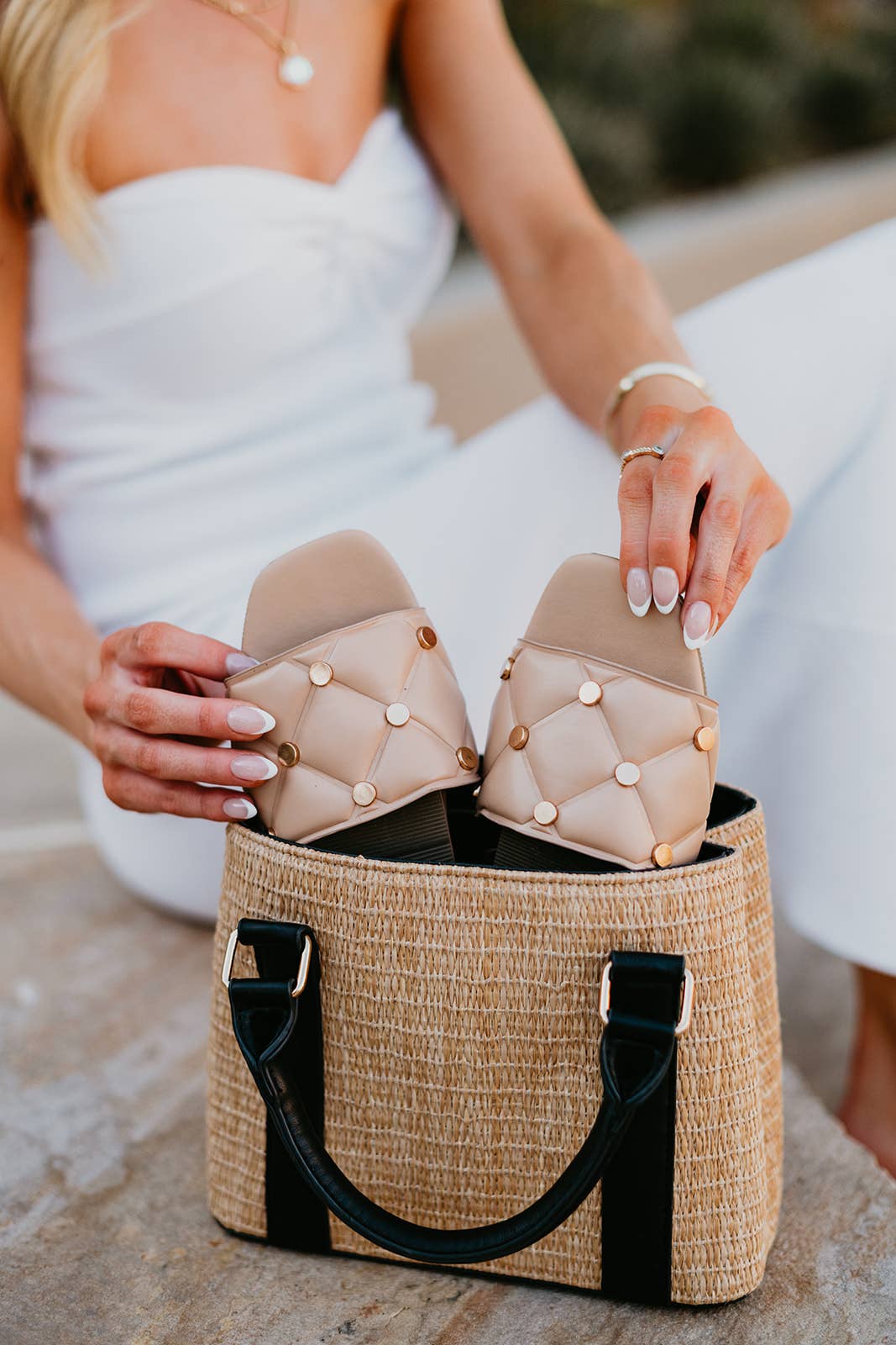 Person holding a pair of beige quilted sandals over a woven bag.