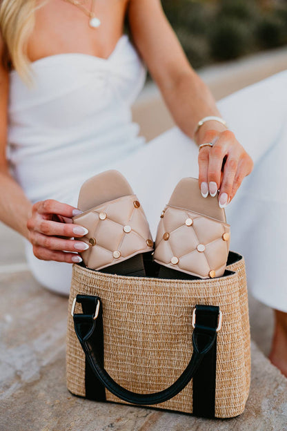 Person holding a pair of beige quilted sandals over a woven bag.