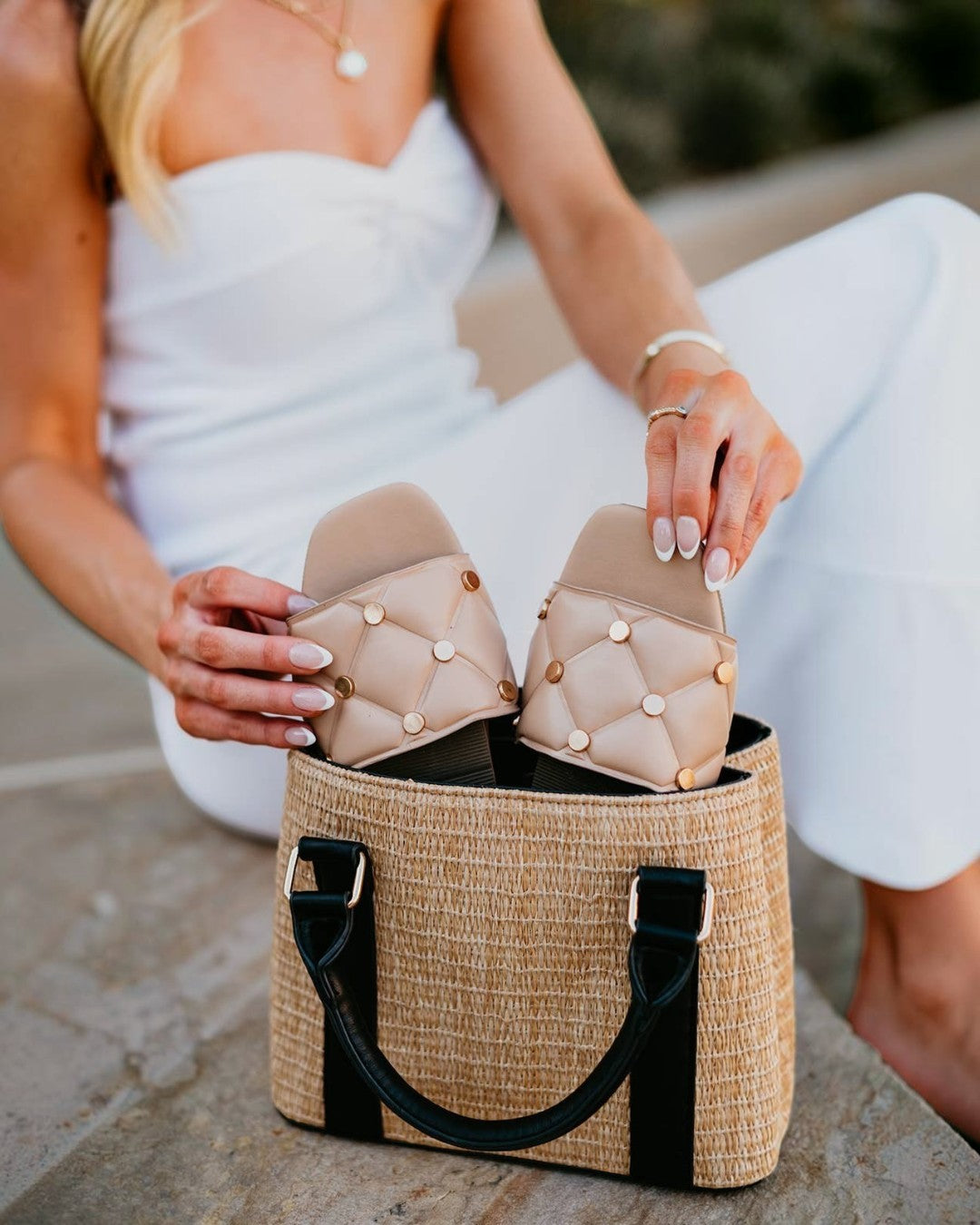 Person holding a pair of beige quilted sandals over a woven bag.