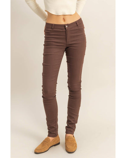 A model wearing brown skinny jeans, styled with a cream long-sleeve crop top and tan suede loafers, shown from the waist down against a light beige studio background.