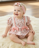 Baby girl in a floral outfit with pink suspenders and headband sitting on a fluffy white rug.