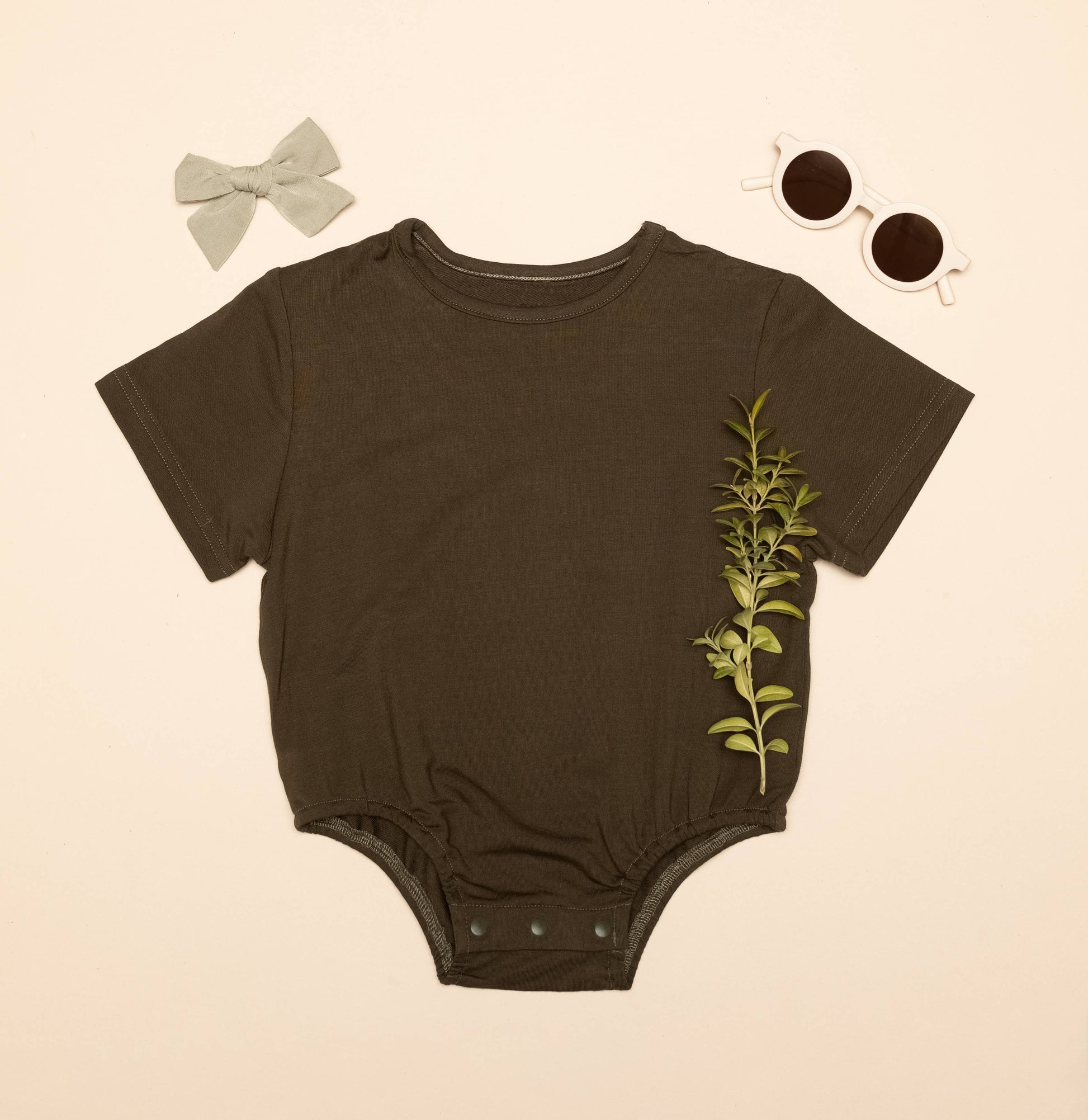 Brown baby onesie with leaf design, gray bow, and sunglasses on beige background