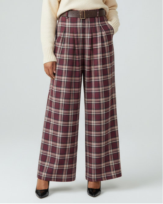 A model stands against a light gray studio background wearing a cream knit sweater, a brown belt, and high-waisted wide-leg plaid trousers in burgundy, beige, and brown. She is wearing black pointed-toe heels.