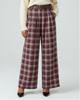 A model stands against a light gray studio background wearing a cream knit sweater, a brown belt, and high-waisted wide-leg plaid trousers in burgundy, beige, and brown. She is wearing black pointed-toe heels.