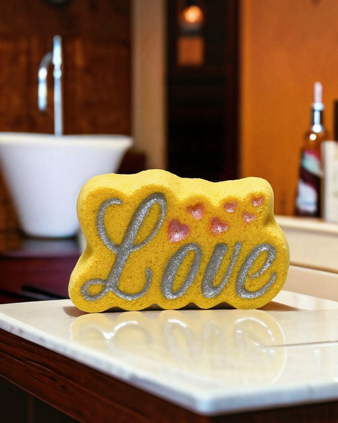 Calla Lily Cosmetics + CL kids yellow Love bath bomb with silver glitter lettering and pink hearts on a marble counter