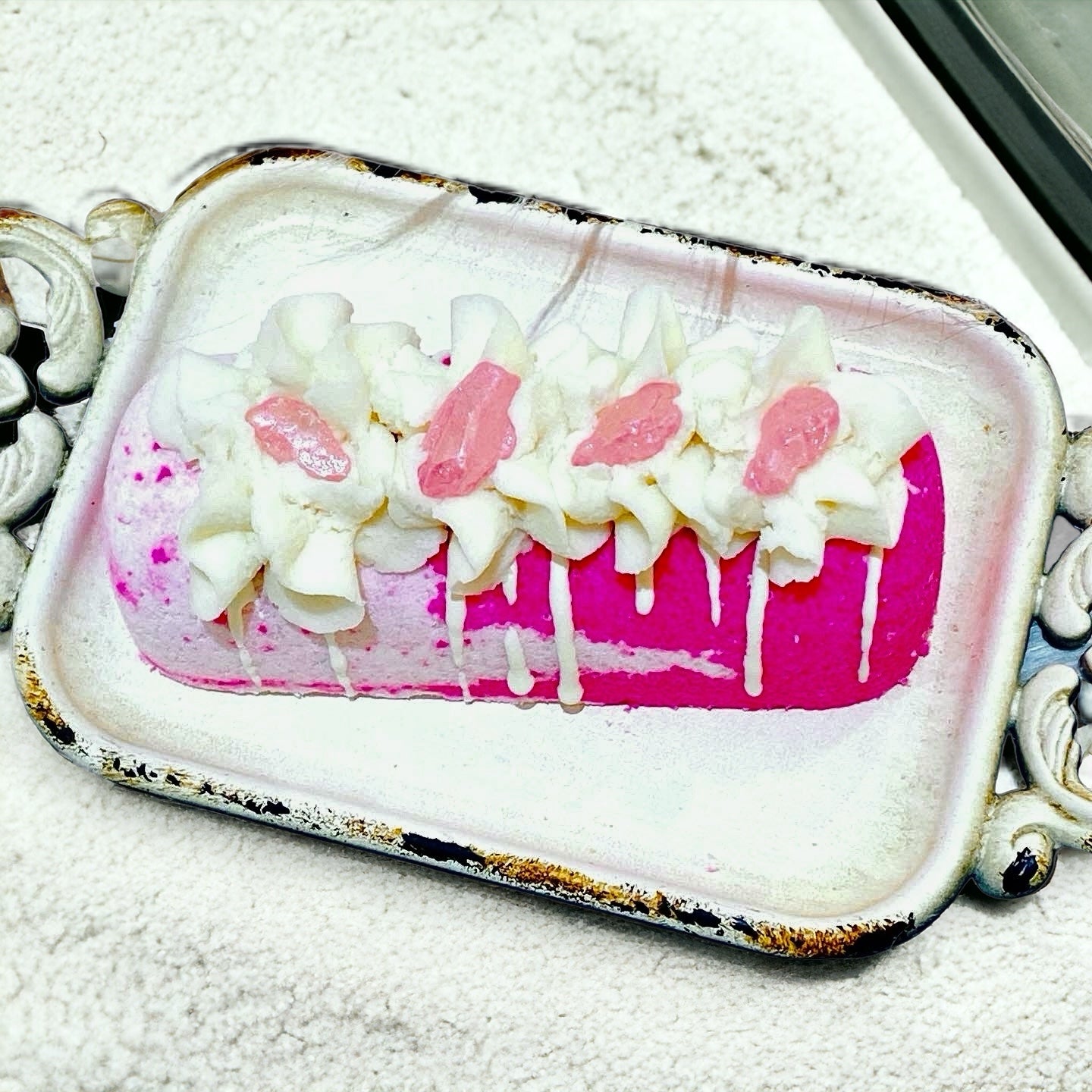 A pink and white twinkie bath bomb topped with white frosting and pink sugar sprinkles on a white tray on top of a white towel