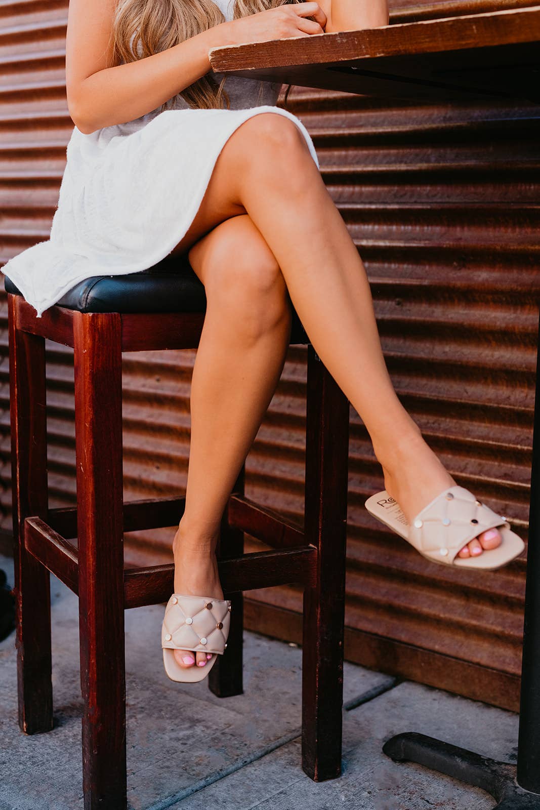 Person sitting on a wooden stool wearing white sandals with a neutral background