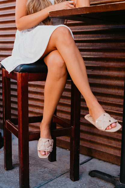 Person sitting on a wooden stool wearing white sandals with a neutral background