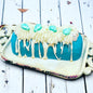 A teal twinkie bath bomb topped with white frosting and teal sugar sprinkles on a white tray against a wood background