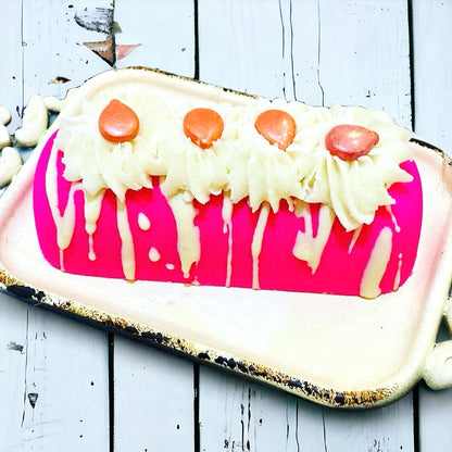 A pink twinkie bath bomb topped with white frosting and sugar sprinkles on a white tray