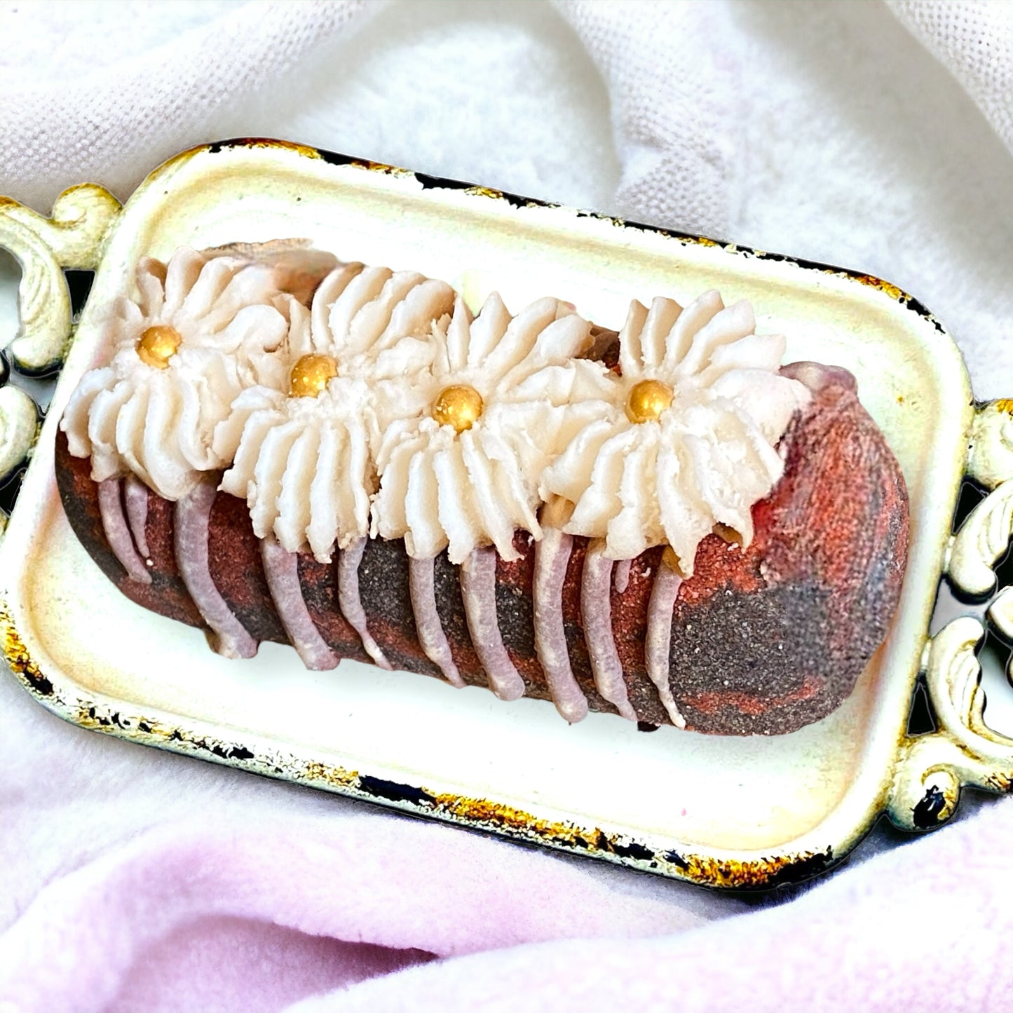 A red and black twinkie bath bomb topped with white frosting and gold sugar sprinkles on a white tray on top of a white towel