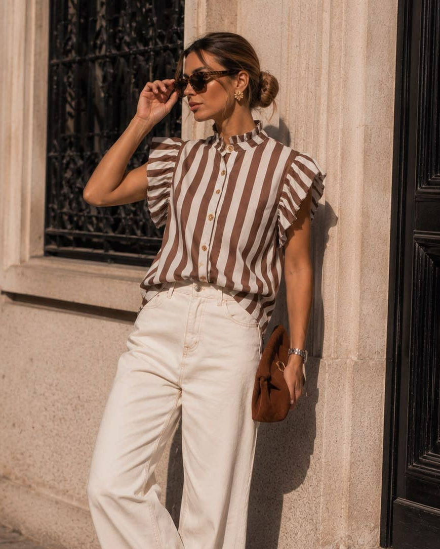A woman stands outdoors against a stone building wall, wearing a brown and beige vertically striped short-sleeve blouse with ruffled shoulders, white wide-leg trousers, brown shoes, sunglasses, and carrying a brown handbag.