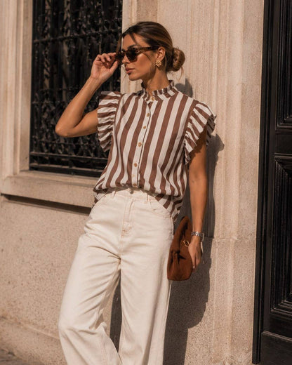 A woman stands outdoors against a stone building wall, wearing a brown and beige vertically striped short-sleeve blouse with ruffled shoulders, white wide-leg trousers, brown shoes, sunglasses, and carrying a brown handbag.