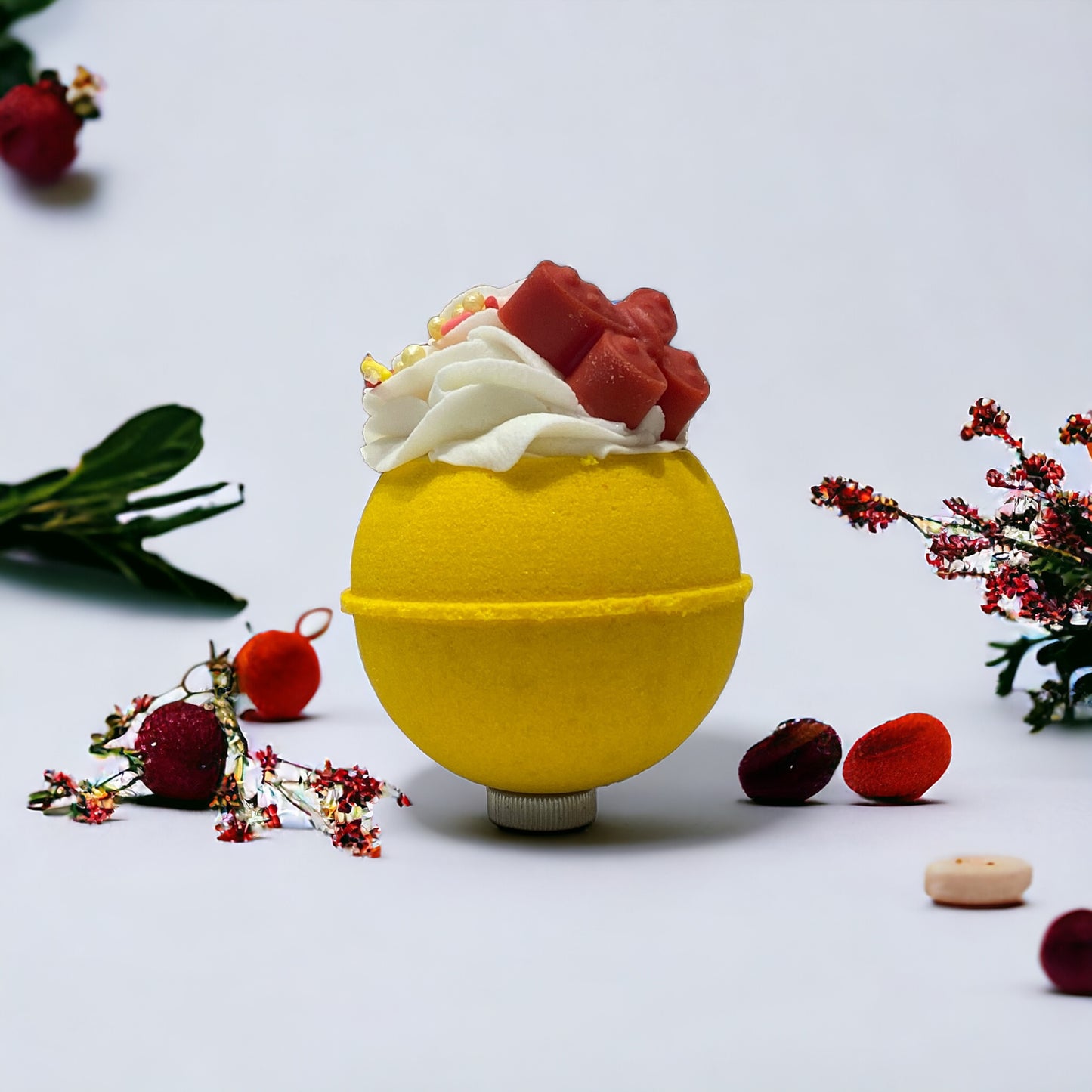 A round yellow bath bomb with white frosting, colorful sprinkles, and a red orange butterfy soap/crayon on top against a white background