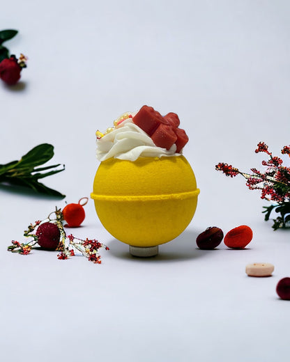 A round yellow bath bomb with white frosting, colorful sprinkles, and a red orange butterfy soap/crayon on top against a white background