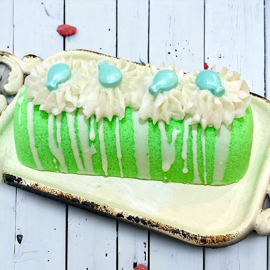 A green twinkie bath bomb topped with white frosting and teal sugar sprinkles on a white tray against a wood background