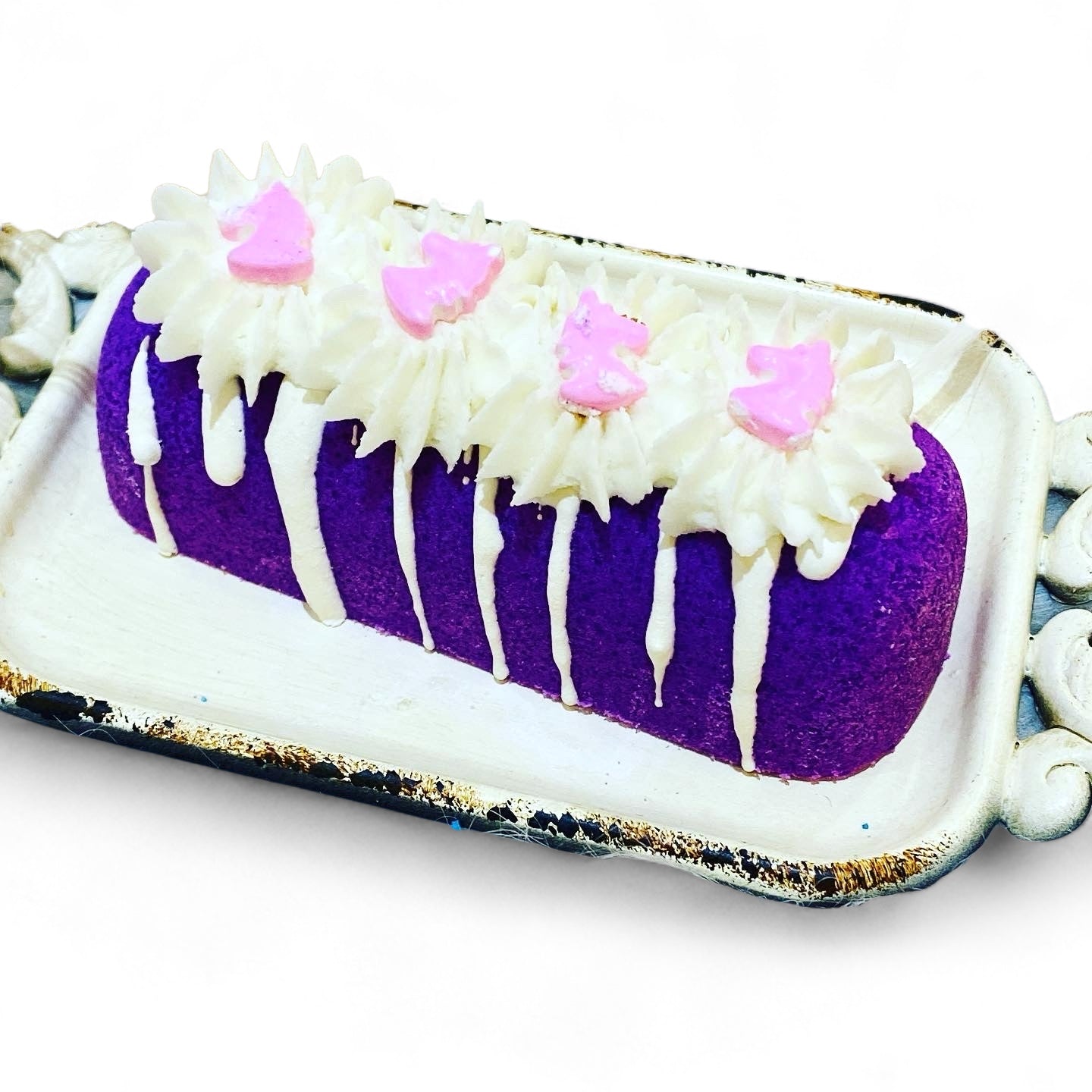 A purple twinkie bath bomb topped with white frosting and light purple sugar sprinkles on a white tray against a white background