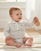 Baby sitting on a rug holding a wooden toy in a bright room.