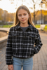 Young girl wearing a plaid jacket and jeans standing outdoors with a blurred background