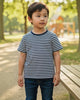 Child wearing a striped shirt standing in a park with trees and a playground in the background