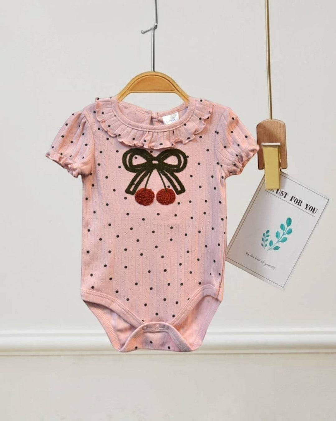 Pink baby bodysuit with black polka dots and bow design hanging on a white wall.