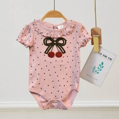 Pink baby bodysuit with black polka dots and bow design hanging on a white wall.