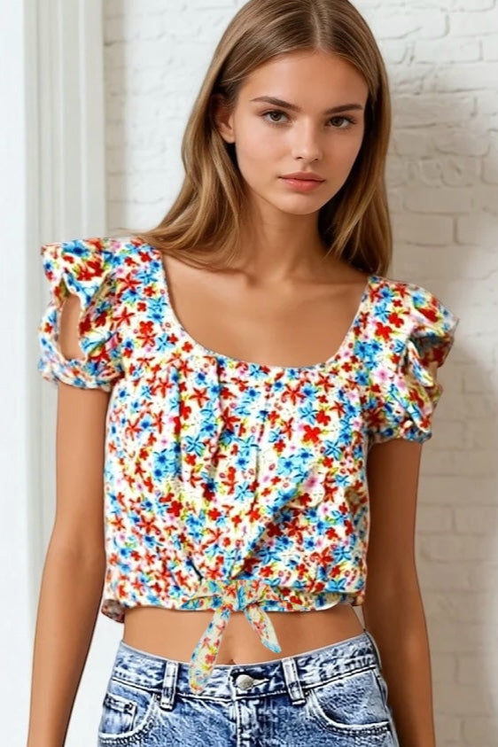 Woman wearing a colorful floral top with ruffled sleeves against a white brick wall.