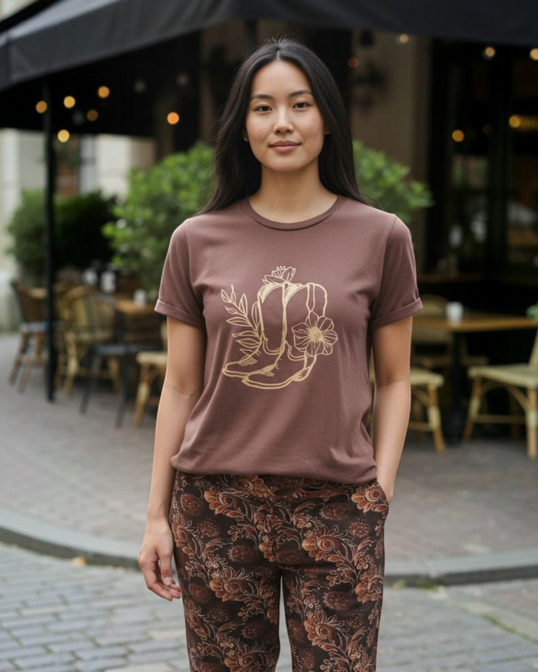 Woman wearing a brown t-shirt with a graphic design and patterned pants standing outdoors.