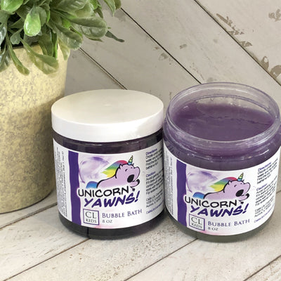 Two jars of 'Unicorn Yawns' bubble bath on a wooden surface with a plant in the background.