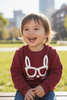 Child wearing a maroon sweatshirt with a white graphic, sitting outdoors with a blurred cityscape in the background.