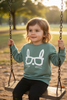 Child on a swing wearing a green sweater with a white graphic, sitting in a park.