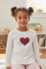 Young girl wearing a white sweater with a red heart design in a room with shelves and books.